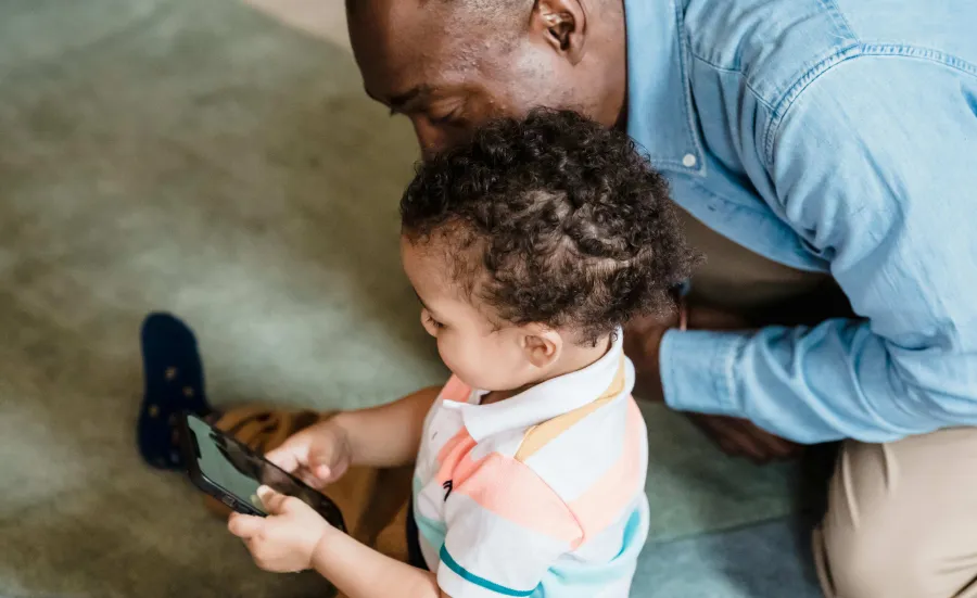 Young child playing on a phone as an adult watches them over their shoulder looks 