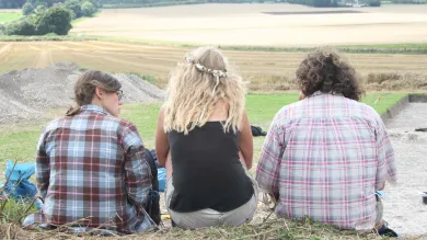 Three people sit on a hill overlooking a large open area of countryside at Avebury, a site of ancient settlement.