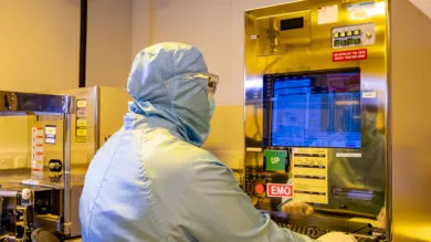 A researcher in a cleanroom suit sits with their back to the camera, using the Critical Dimension Scanning Electron Microscope (CD-SEM) 