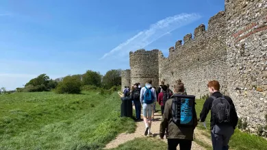A group of history students walk beside the outside wall of the Hampshire medieval fortress Portchester Castle, as part of a field trip. The 2 students closest to us are in discussion.