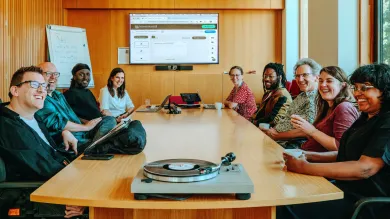  A group of nine people is seated around a wooden conference table, smiling and engaged in a meeting. Laptops are open, and a turntable with a vinyl record sits at the front of the table. A large screen behind them shows a web interface, and a whiteboard with notes is visible. The mood is friendly and collaborative in a modern, well-lit room.