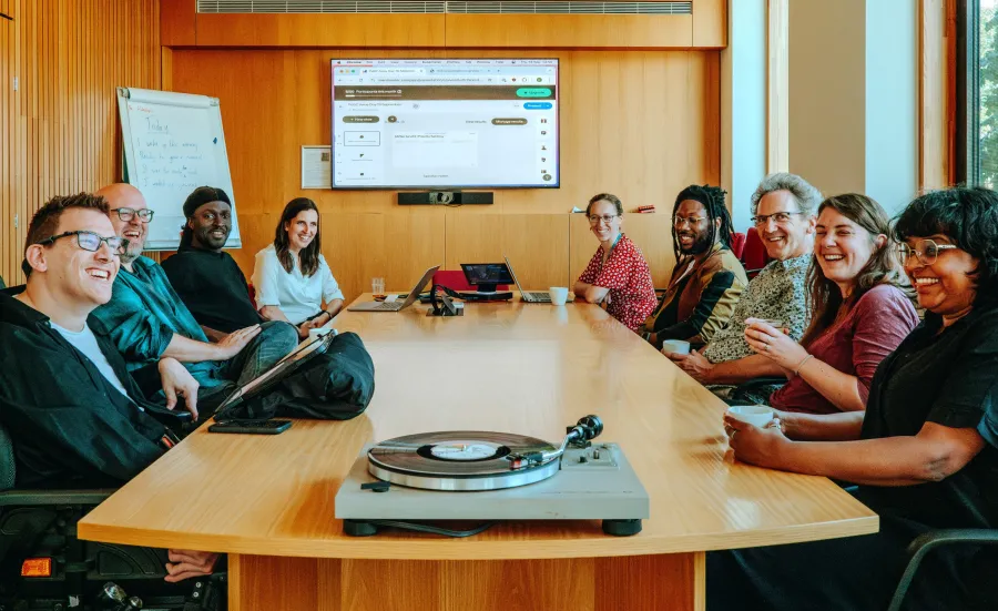 A group of nine people is seated around a wooden conference table, smiling and engaged in a meeting. Laptops are open, and a turntable with a vinyl record sits at the front of the table. A large screen behind them shows a web interface, and a whiteboard with notes is visible. The mood is friendly and collaborative in a modern, well-lit room.