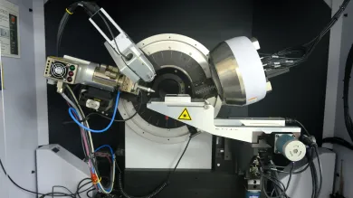 Complex scientific instrument, likely an X-ray diffractometer, set up inside a laboratory environment. It features a cylindrical component at the centre, surrounded by various metallic parts and wiring. 