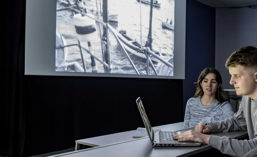 Two film students looking at a laptop in a lecture theatre. There is a film showing on a laptop and there is a large screen behind them showing a film of small boats.