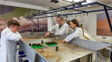 Three people in lab coats work at a sand-filled table with various objects and overhead nozzles, set against a backdrop of landscape images.