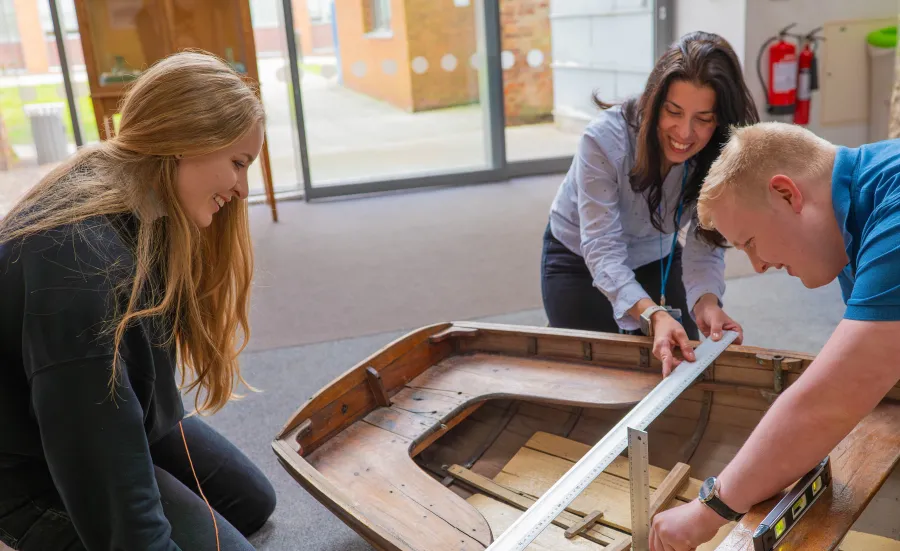 Ancient History students and professor measuring a replica wooden boat with a metal ruler. The boat is on the floor of a university building and the people are kneeling around the boat.