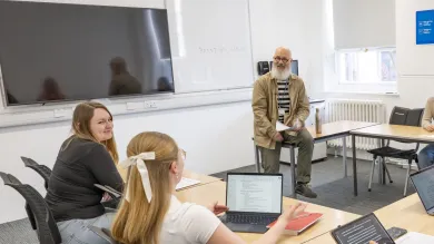 Alt text: A teacher sits on a desk holding papers while three students face them, one using a laptop. A whiteboard behind the teacher displays the phrase "POINT OF VIEW."