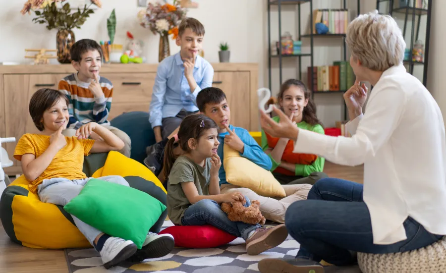 A group of primary school age children are sitting on bean bags on the floor. They are engaged watching a speech therapist talking to them and showing them a large letter ‘S’. They are all touching their chin with an index finger.