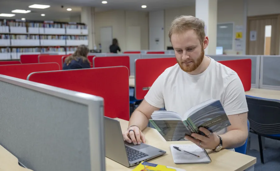 Public Health student, Jack Coles working at a desk in the library. He is reading a book and typing on a laptop keyboard.  