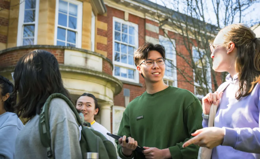 Two postgraduate students smiling at each other while walking outside the Parkes Building, also known as Building 65, on the University of Southampton's Avenue Campus. They are part of a larger group of students.