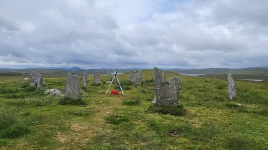 A grassy field features a circle of weathered standing stones, some covered in lichen. A tripod and red bag are positioned near the center under a cloudy sky.