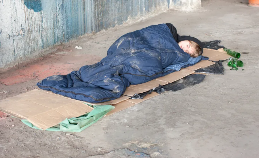 Image shows someone sleeping rough. They're sleeping on the streets in a blue sleeping bag on top of some cardboard.