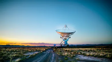 A towering satellite dish positioned in a wide, grassy field, silhouetted against a vivid orange sunset sky, with a winding dirt road leading toward it.