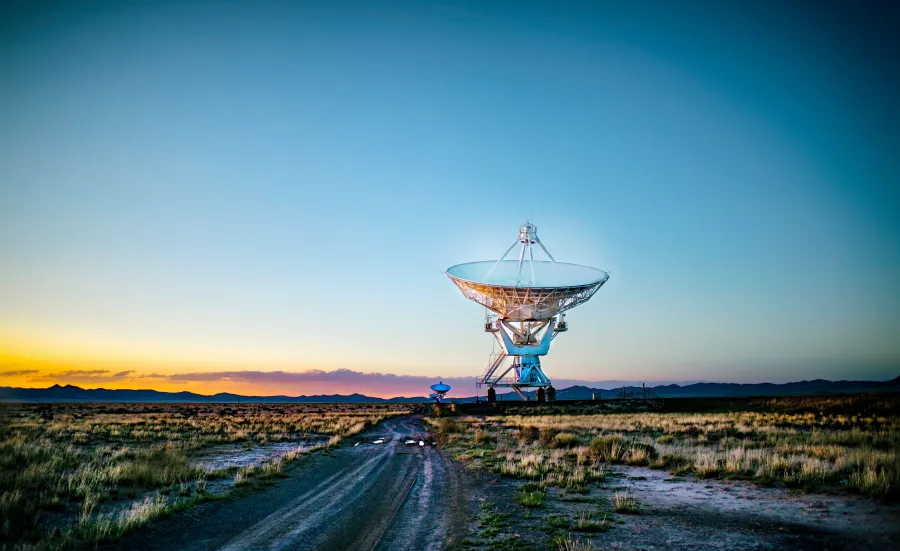 A towering satellite dish positioned in a wide, grassy field, silhouetted against a vivid orange sunset sky, with a winding dirt road leading toward it.