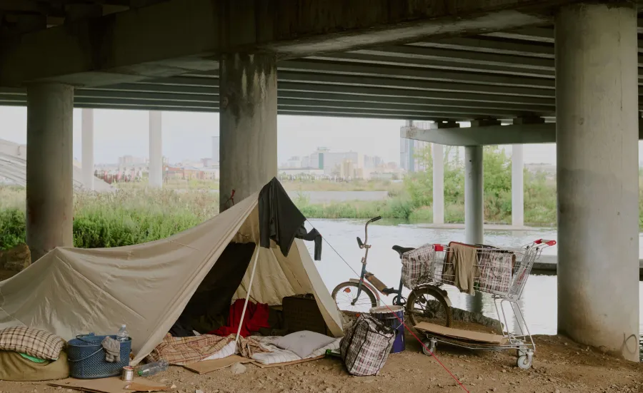 A makeshift shelter set up under a bridge near a river, with personal belongings, a bicycle, and a shopping trolley. The image reflects urban homelessness and temporary living conditions