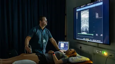 A medical professional uses an ultrasound device on a woman lying on an examination table, with a large screen displaying the ultrasound image.