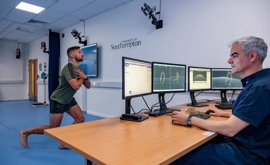 A man performs a lunging squat with motion capture markers, while another man monitors data on screens in a lab.