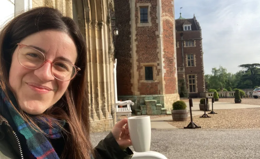 A image of Chiara smiling at the camera. She's sat down outside and is holding a white mug. There's a brick building and grounds behind here.