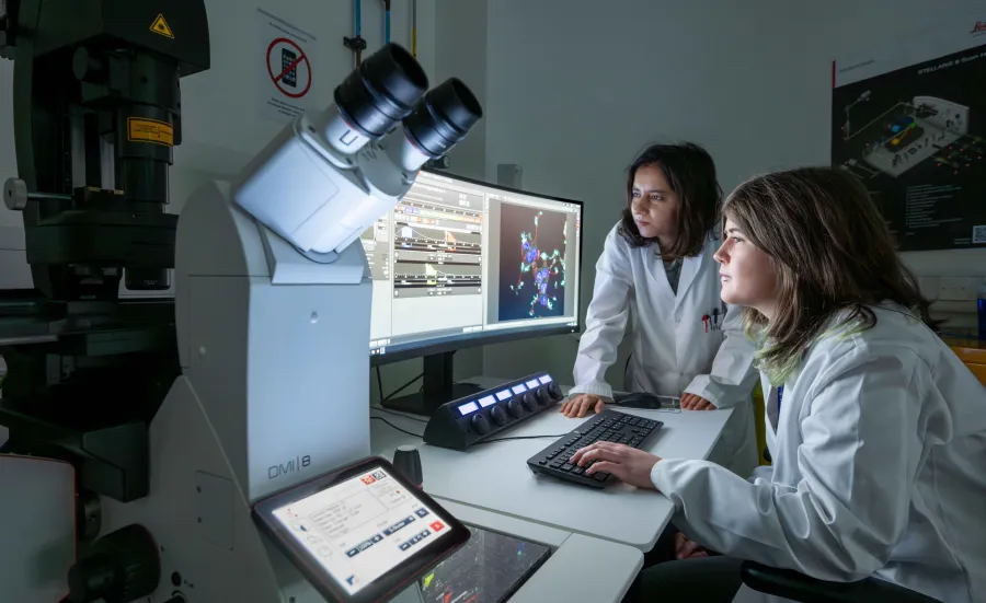 Two students in white lab coats using the microscopy lab facility. They're both analysing data on a computer monitor and there's a large microscope to the side of them.