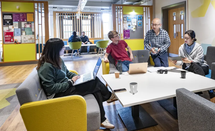 A group of postgraduate students working and chatting together in a modern campus social space.