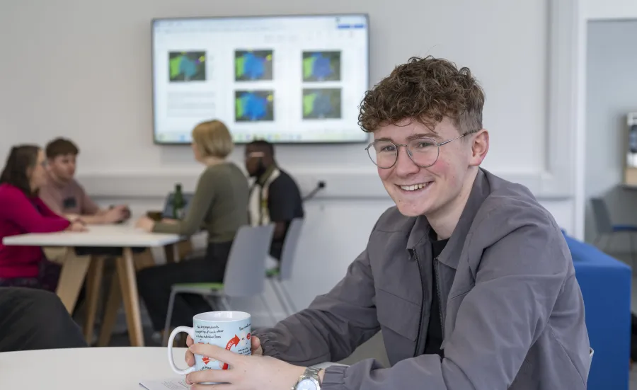 Samuel sitting at a table in a communal area. He has a mug in his hands and is smiling for the camera. 