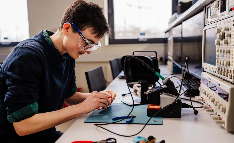 Biomedical engineering PhD student wearing safety goggles soldering a circuit board in an electronics lab.