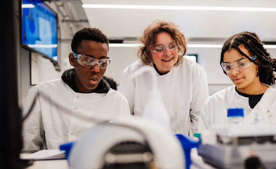 Three students in lab coats and safety goggles working together in a science laboratory.