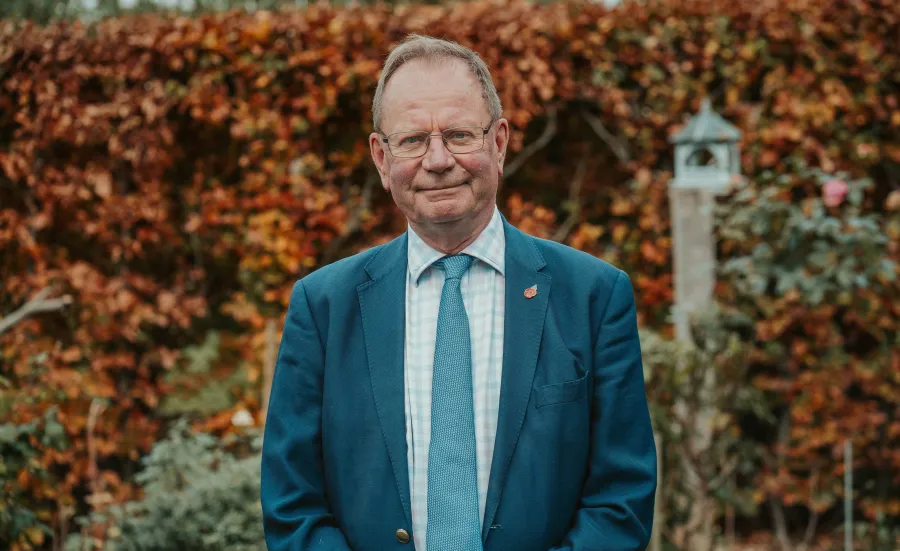 Portrait of Professor Stephen Holgate in a blue suit standing outdoors in front of autumn foliage.