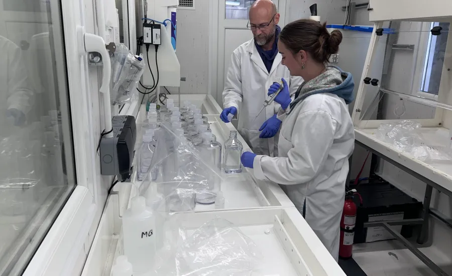 Two people in a laboratory preparing water samples, wearing lab coats and gloves, and handling bottles and equipment on a long workbench.