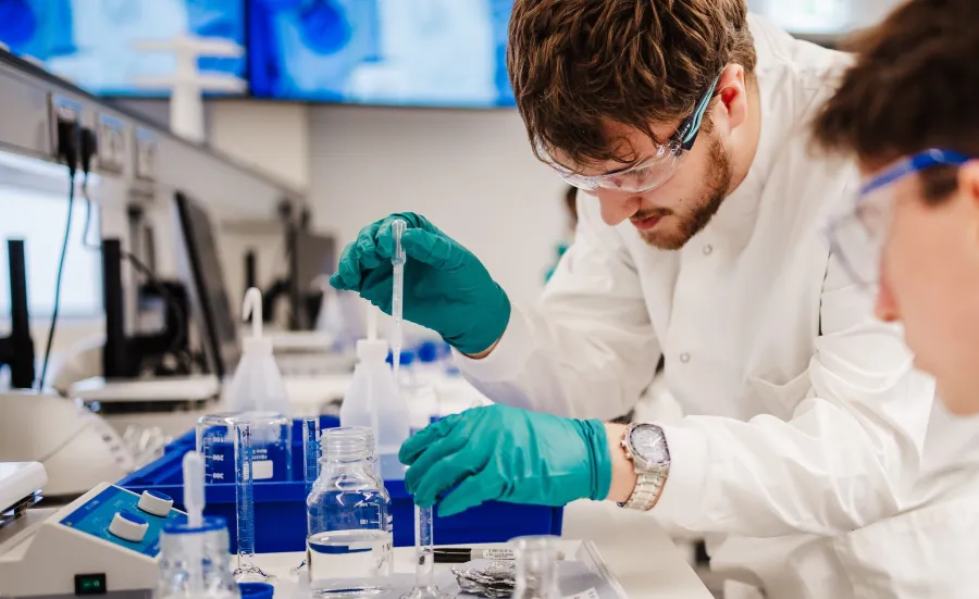 Students wearing safety goggles and gloves conduct a laboratory experiment using pipettes and glassware.