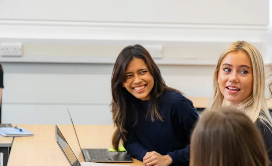 A group of students smiling having a discussion. There are open laptops on the table in front of them.