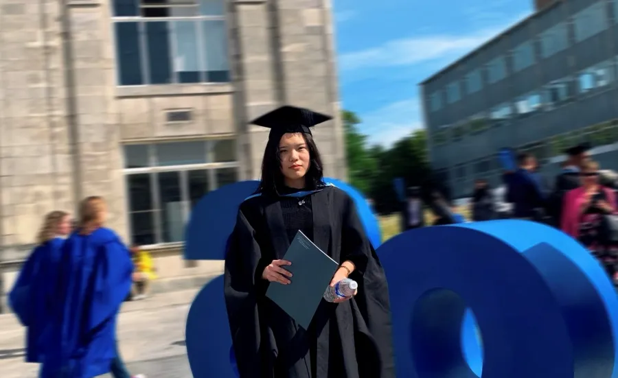 A person wearing graduation robes and a mortarboard stands outdoors holding a certificate folder, with large blue sculpture letters and university buildings in the background.