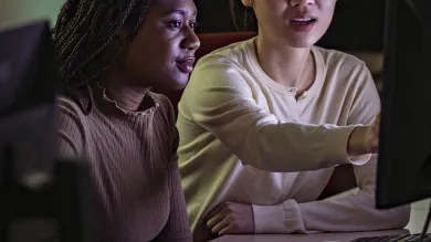 Two students seated together in the Bloomberg suite. One student gestures towards something on a computer screen.