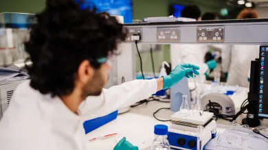 A biomedical engineering student measuring a solution with a thermometer. The student is wearing a white coat and eye protection.