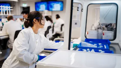 A biomedical engineering student adding a crate of samples to a fume cupboard. The student is werring a white coat, eye protection and gloves.
