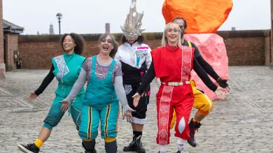 Five performers in bright costumes walk towards the camera at the British Science Festival 2025 in Liverpool. There is a large orange sculpture behind them.