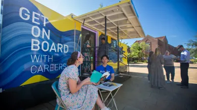 Students speaking and reading outside a careers bus on campus, with others queuing and talking nearby.