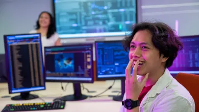 A student turns to the camera in a lab. He has a hand to his mouth and looks pleasantly surprised. Several monitors display different sets of data and another student is seen in the background, curious as to what has surprised him.