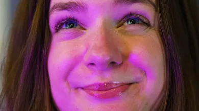 The camera leans close to the face of a student bathed in a warm purple light. She looks up, smiling expectantly