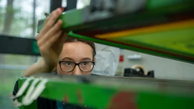A person wearing glasses adjusts or inspects equipment in a laboratory or workshop setting, viewed through green-tinted panels. The image focuses on close observation and hands-on technical work.