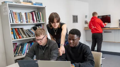 Three students working collaboratively with laptops at a desk in a university study space.