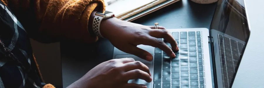 Fingers typing on a laptop with notepad, pen and mug in the background