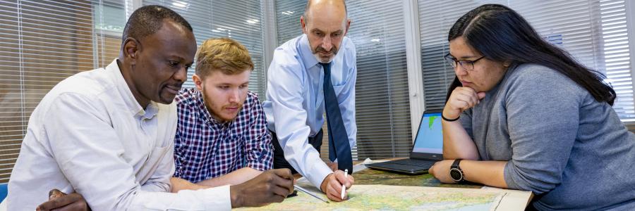 A Southampton Geospatial researcher talks with team members, standing over a map, with a laptop with a data visualisation to the side