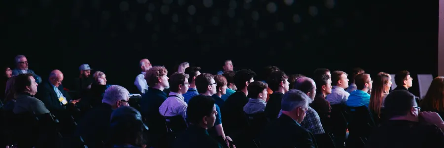 A three quarter view from behind of conference attendees sat in rows. The room is mostly dark and there is light coming from the front of the room, out of the shot. 