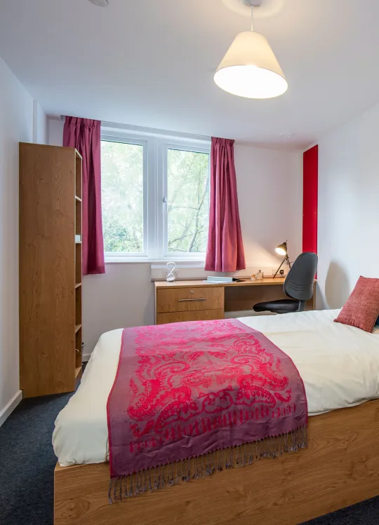 Bedroom with a neatly made double bed and nightstand. A wooden desk and open window are visible in the background.