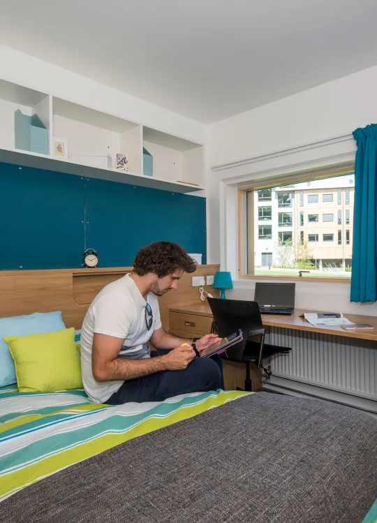 A student sits using a tablet on a large, brightly coloured bed in a modern bedroom. Modern buildings and a grassy courtyard can be seen through the windows.
