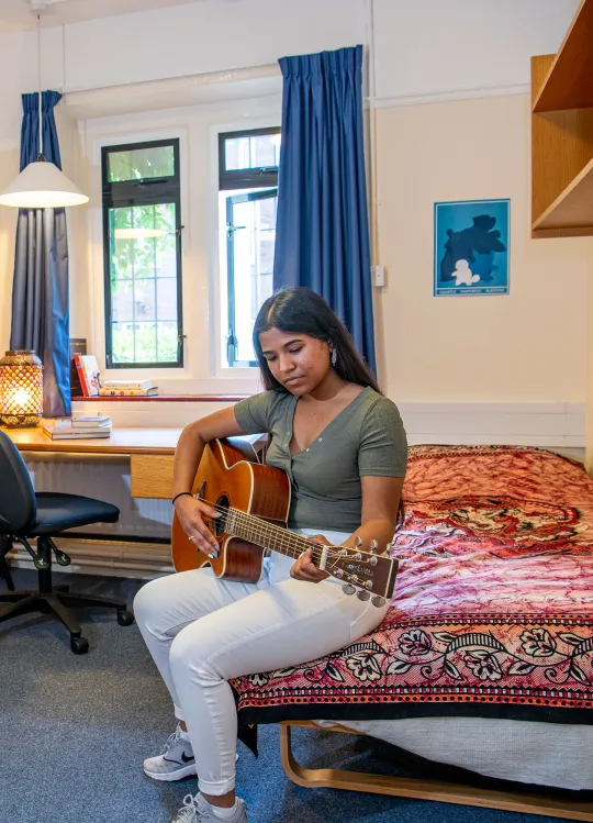 A student plays a guitar while sitting on a bed in a bedroom. In the background, shelves and a desk with books can be seen.