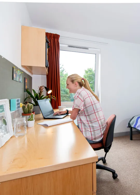 A spacious, bright student bedroom showing a single bed, desk and open window. A student sits at the desk, working on a laptop.