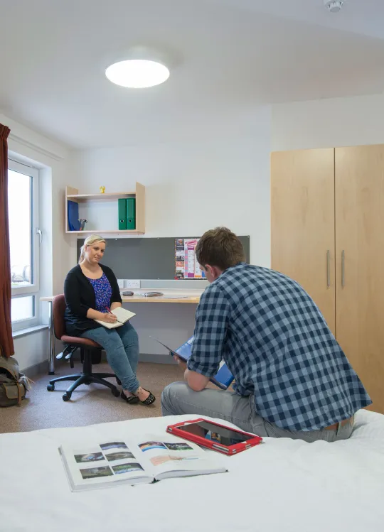 Two students sat talking in a large spacious bedroom. A double bed, desk and open door to a bathroom can be seen.
