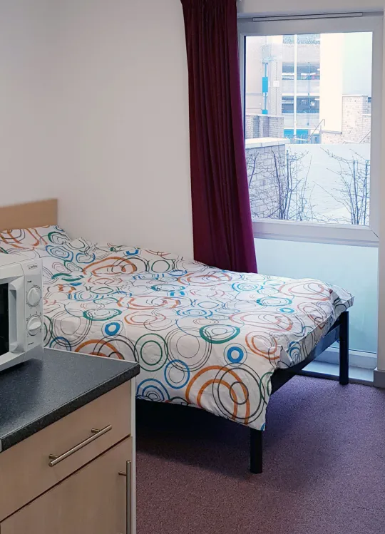 View of a studio flat showing a double bed and wooden desk. A kitchen area with microwave and hob is also partially visible.